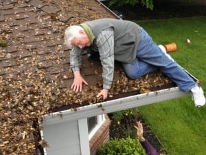 Person sitting on a shingle roof reaching down to hand-clear leaves out of a packed gutter