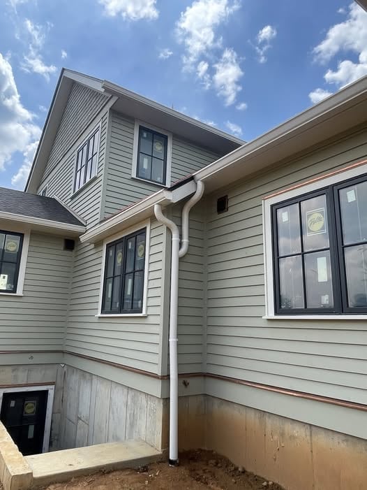 Newly installed white K-style gutter and downspout running down a sage-green clapboard wall on a new-construction home